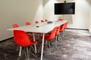 Konferensrum med vitt bord, röda stolar, konferenstelefon och väggmonterad skärm