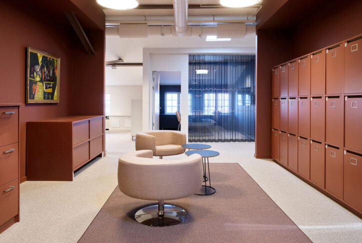 Kontorsyta med beige stolar, rött skåp, tavla, skåpvägg och stora fönster.