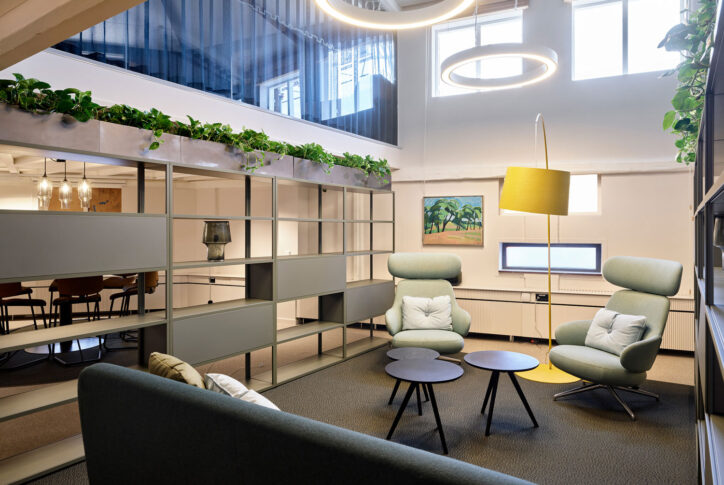 Kontorslounge med fåtöljer, soffor, bord, växter, glasvägg och mötesrum i bakgrunden.