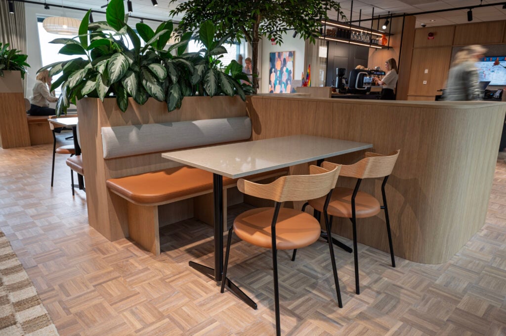 Lunchutrymme med träbord, stolar och bänk, omgiven av gröna växter.