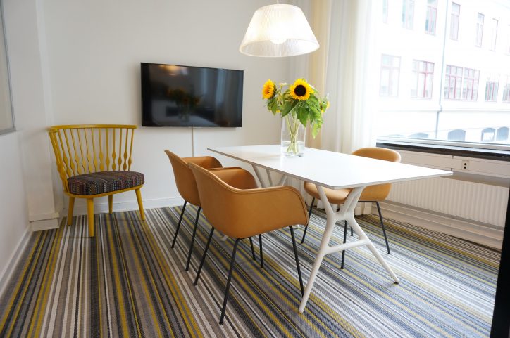 Ljus rum med vitt bord, bruna stolar, solrosor, gul stol, TV och randig matta.