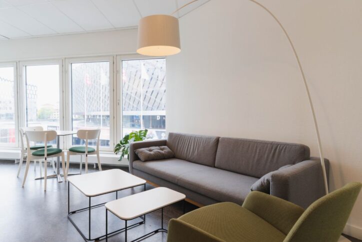 Minimalistiskt lounge med grå soffa, gröna stolar, vita bord och stora fönster.