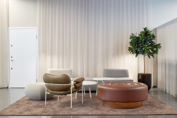 Lounge med brun sittpuff, grå fåtöljer, växt och ljusa gardiner.