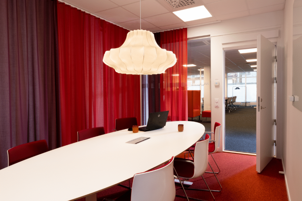 Konferensrum med vitt bord, röda stolar och hängande lampa.