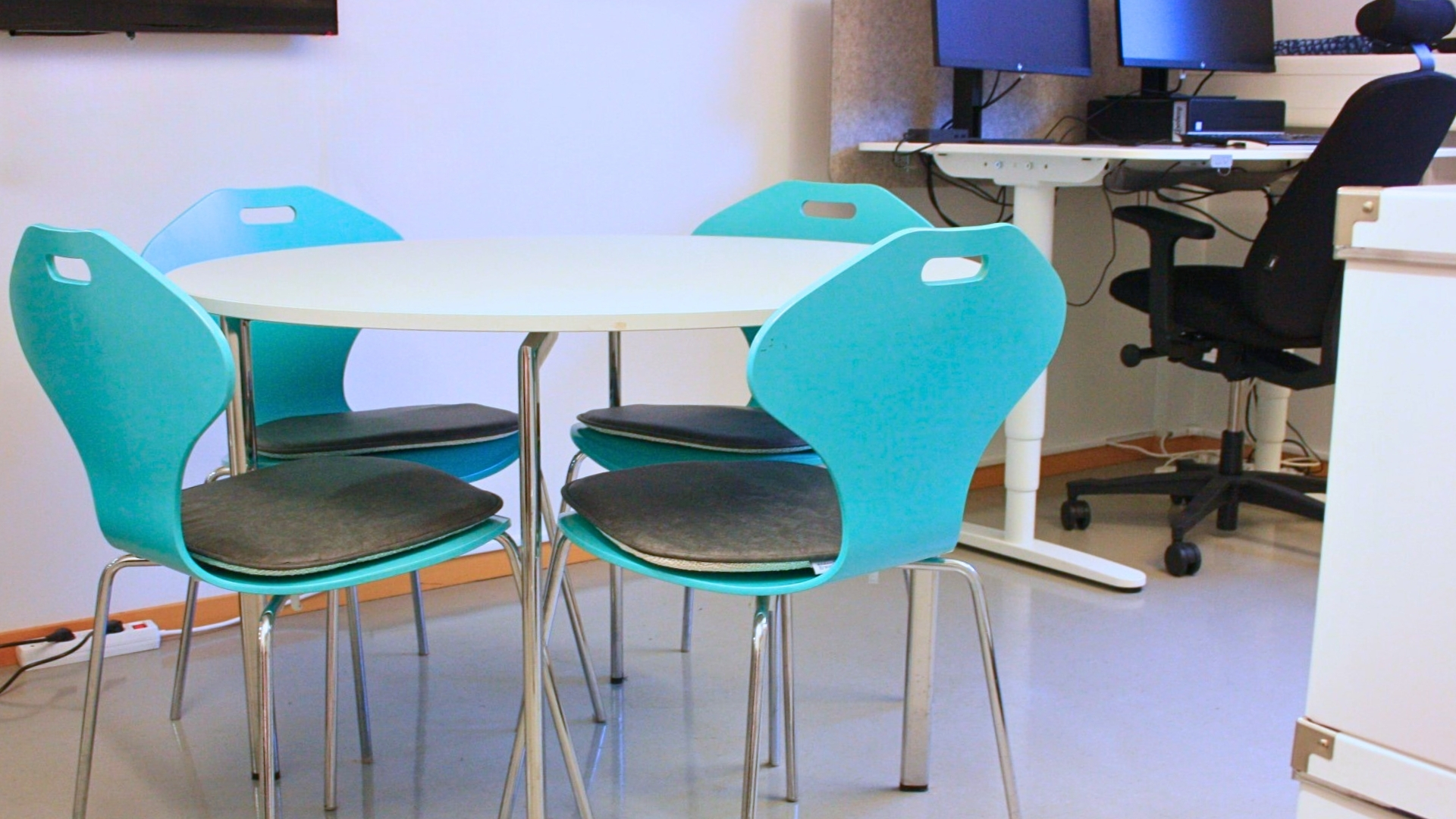Litet mötesrum med rund bord, turkosa stolar och arbetsplats med två datorskärmar.