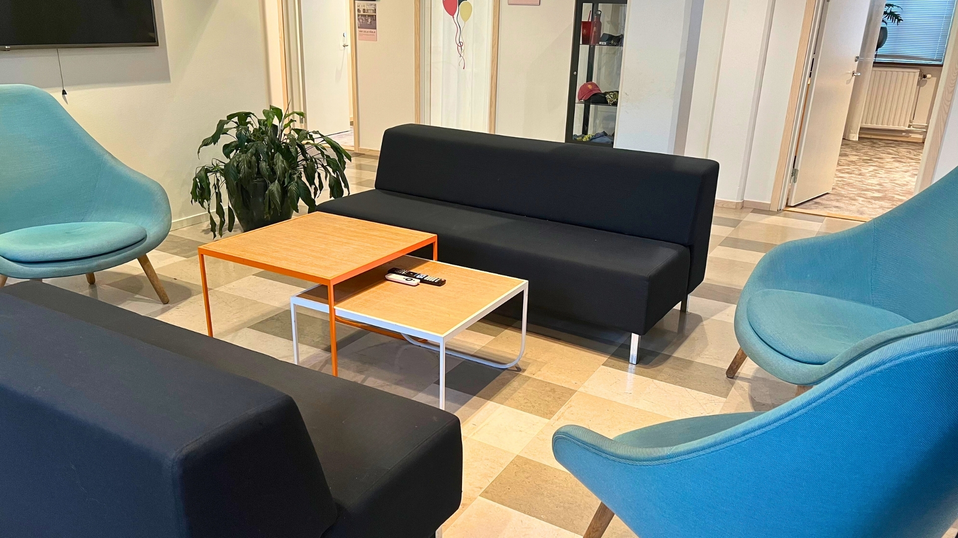 Lounge med soffor, färgglada bord, blå fåtöljer och växt i hörn av kontorsmiljö.