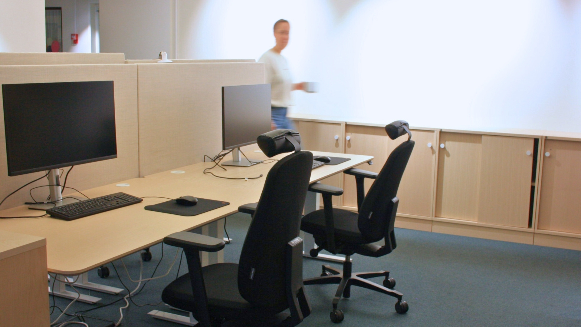 Två arbetsplatser med datorer och stolar i ett modernt kontor med förvaring i bakgrunden.