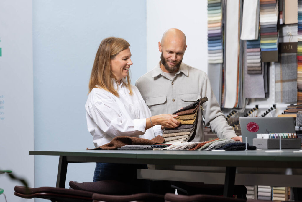 Två personer står vid ett bord och tittar på tygprover, omgivna av färg- och materialprover.
