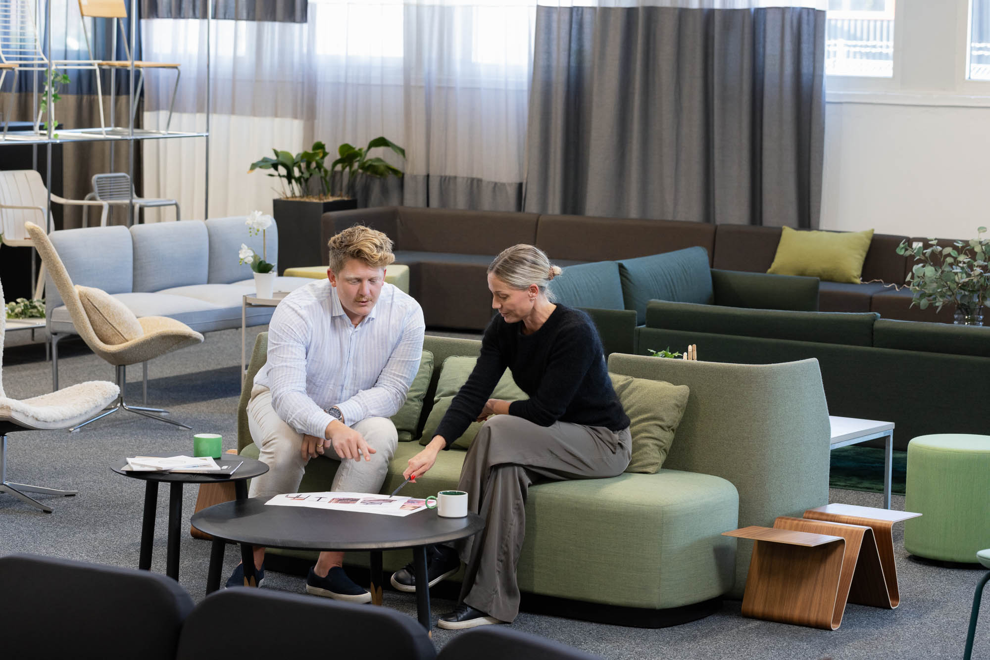 Modern lounge med tre personer som samtalar, soffor i gröna toner, bord med växt och koppar.
