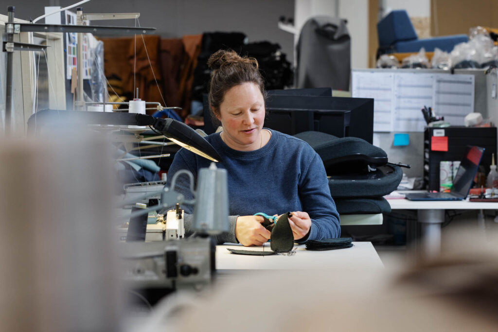 Person arbetar vid ett bord med textil och verktyg i en verkstadsmiljö med maskiner och material.