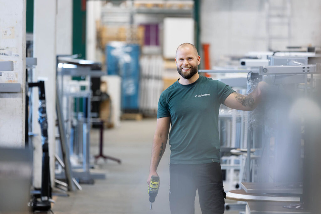Person håller en skruvdragare och arbetar bland metallramar i en industriell verkstadsmiljö.