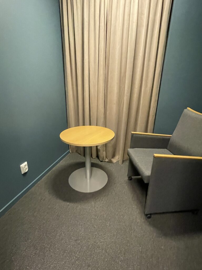 Avskilt rum med fåtölj, litet bord och gardiner mot blå vägg.
