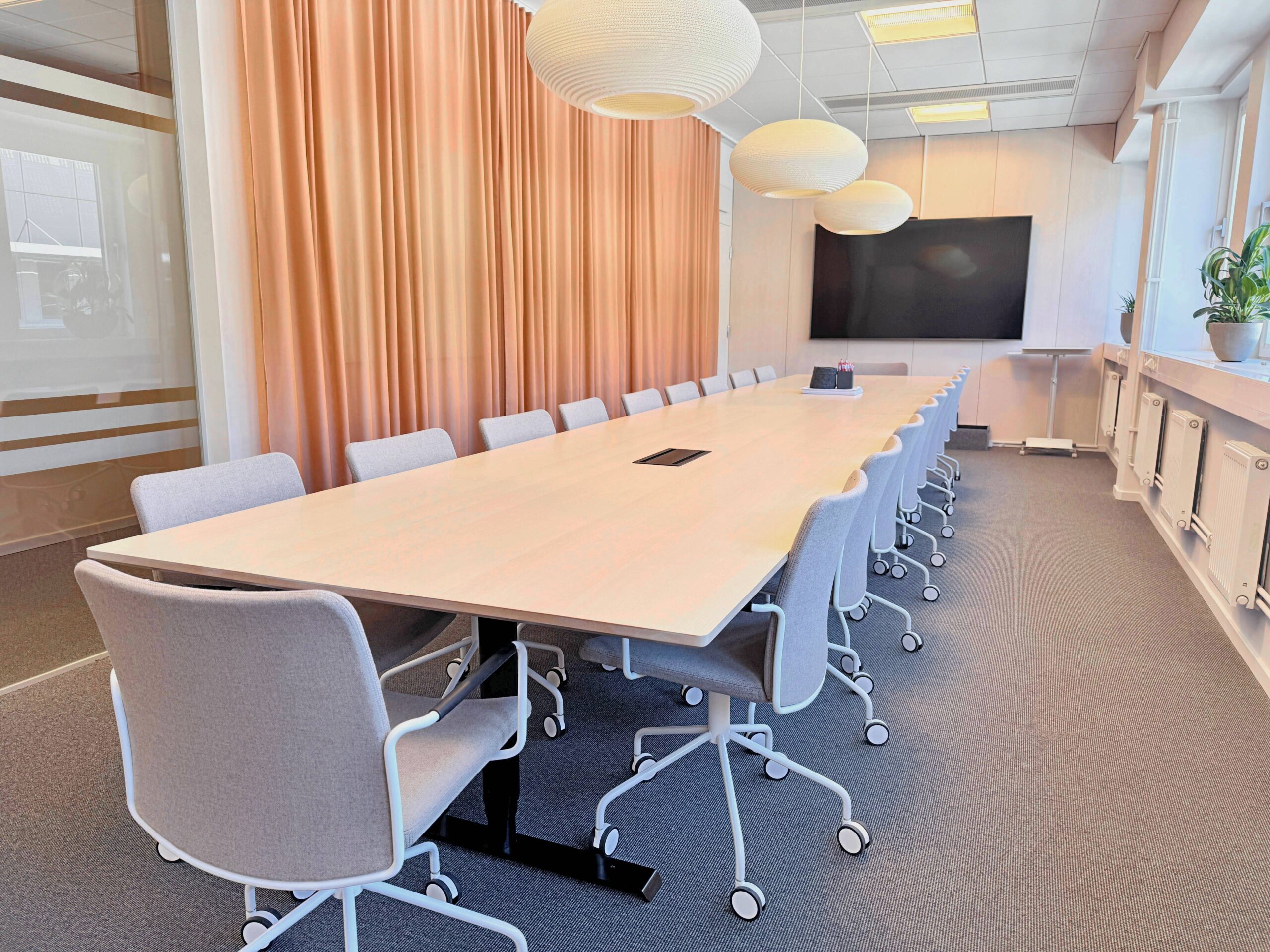 Stort konferensrum med långt bord, stolar på hjul, gardiner och väggmonterad skärm