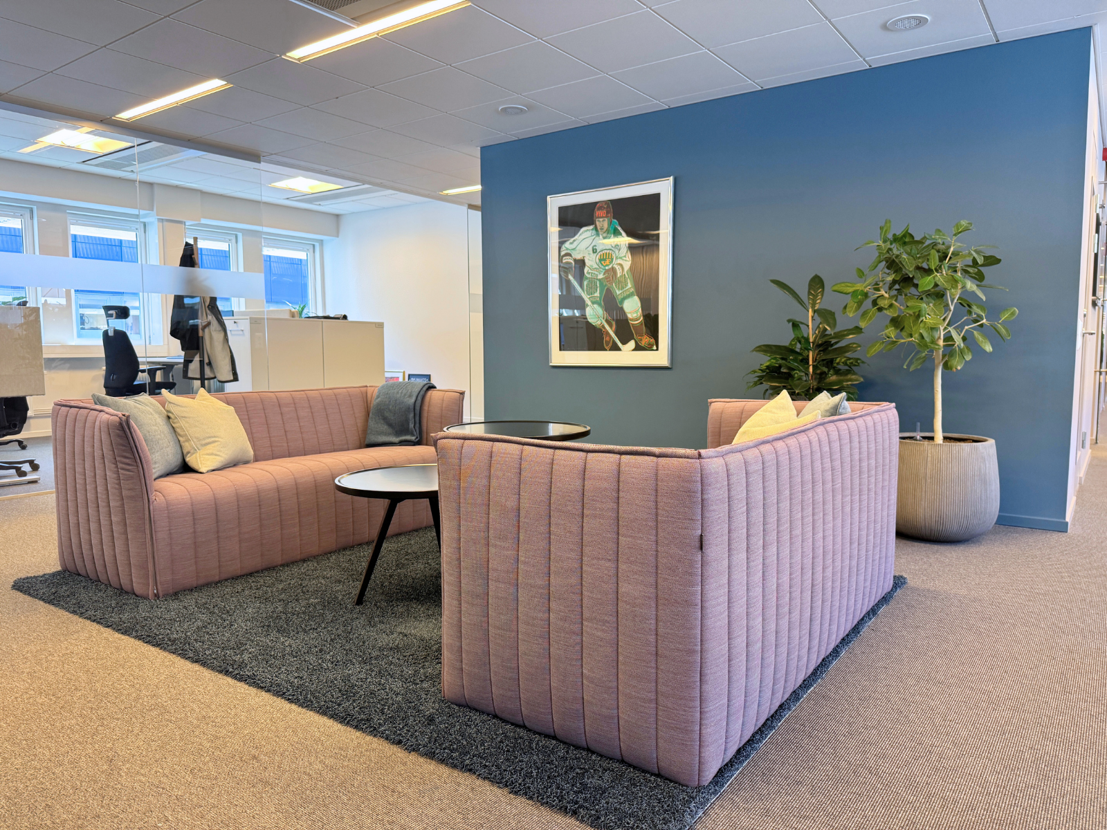 Lounge med rosa soffor, småbord, matta, växt och glasvägg till öppet kontorslandskap