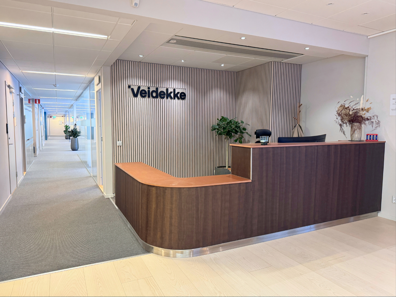 Reception i modernt kontor med trädisk, Veidekke-skylt och ljus korridor med glasväggar
