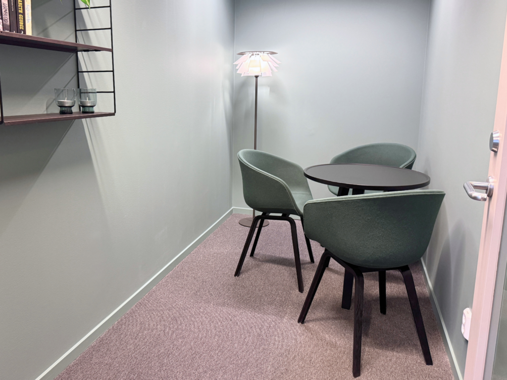 Runt mörkt bord med tre gröna fåtöljer och en golvlampa i ett litet samtalsrum.