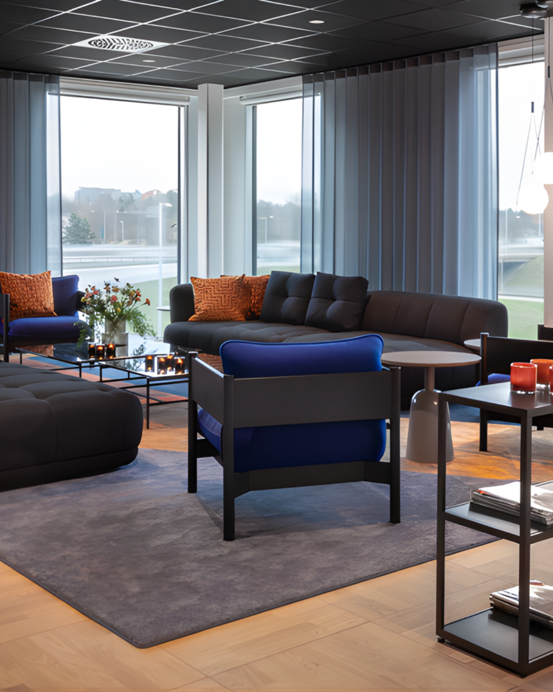 Loungedel med en mörk soffa och fåtöljer placerade på en matta framför fönster med gardiner.